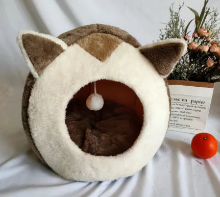 Cat-shaped pet bed with a small white ball inside, placed on a light surface with a plant and paper in the background.